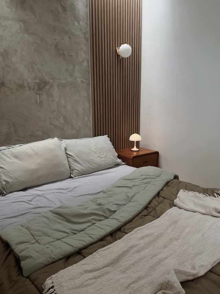 Modern bedroom with wood tone and exposed cement walls, featuring a nightstand and table lamp creating a warm atmosphere