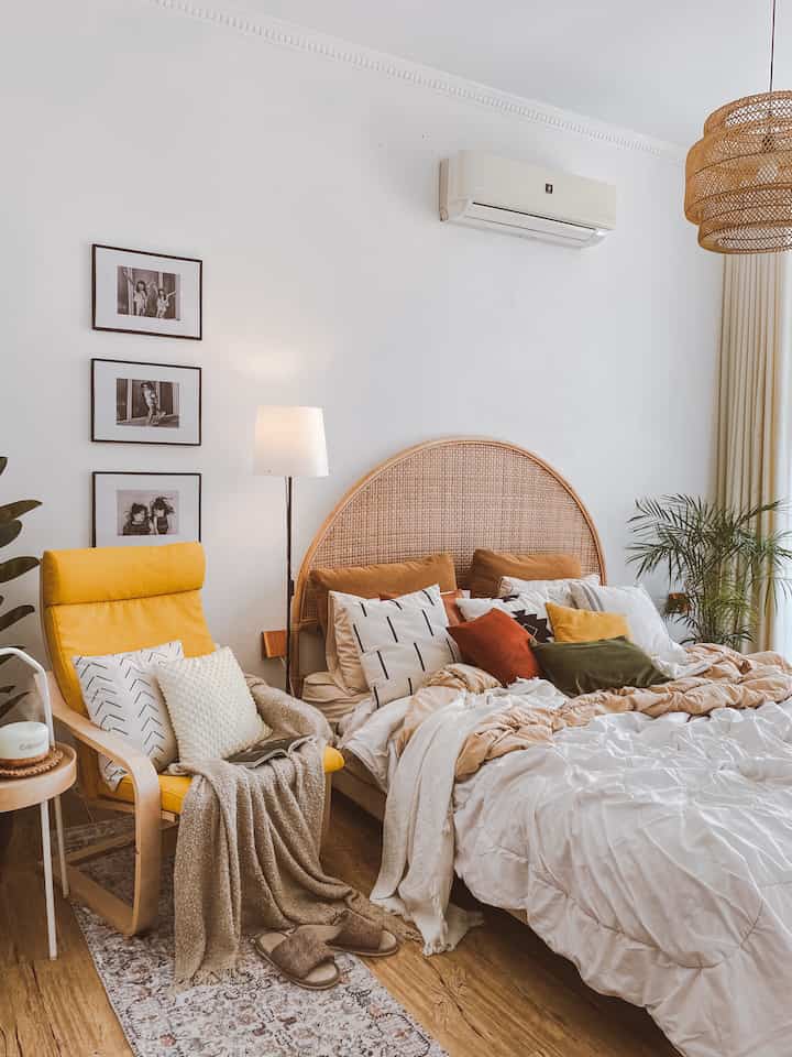 A white and wood tone boho-style bedroom featuring a rattan arch headboard and a yellow armchair creating a cozy atmosphere