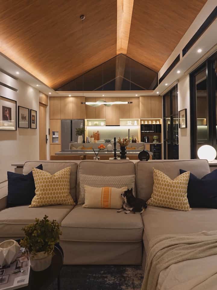 Modern living room with natural wood tones and beige sofa, featuring a pet dog on the sofa and warm cozy lighting