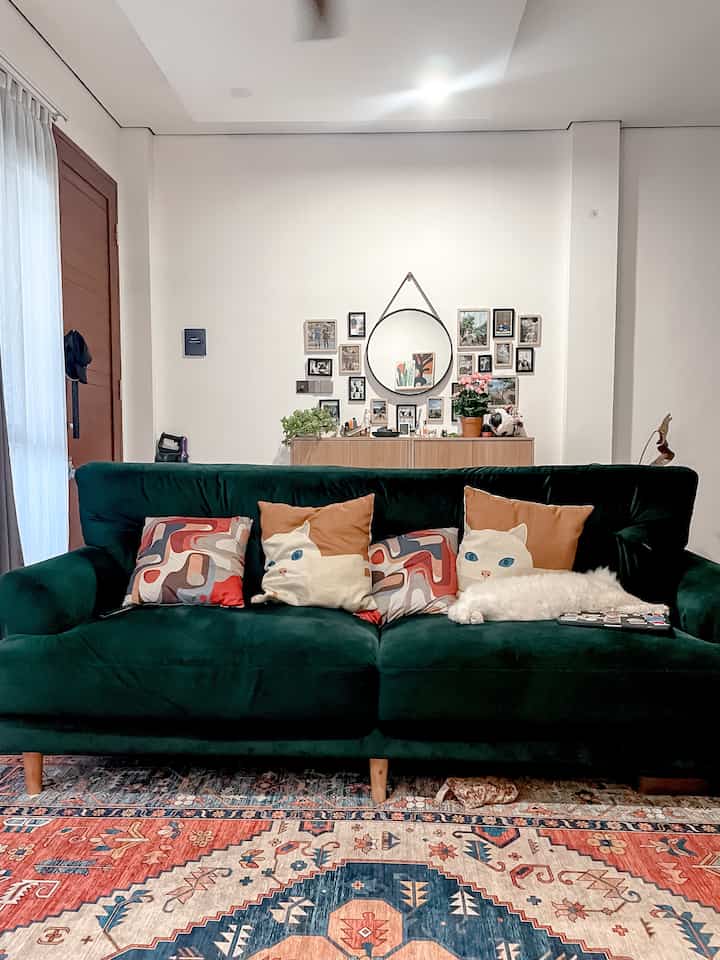 A cozy living room featuring a dark green sofa with patterned cushions, colorful rug, and decorative wall art with mirror and photos