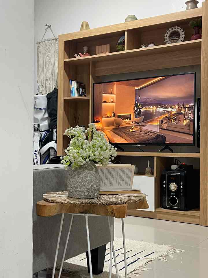 Light brown wood tone living room featuring a gray sofa, TV cabinet, and a wooden side table with flowers on a plush floor rug in a simple modern setting