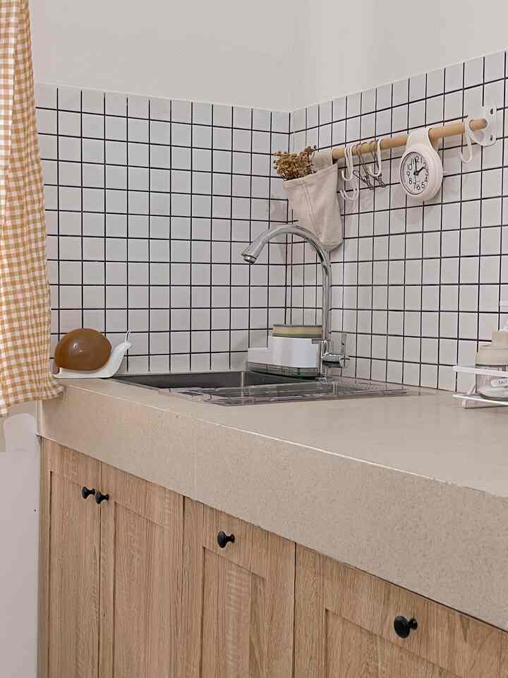 White and wood tone kitchen featuring a clean sink area, grid pattern tiles, gingham curtain, and wall clock in a simple setting
