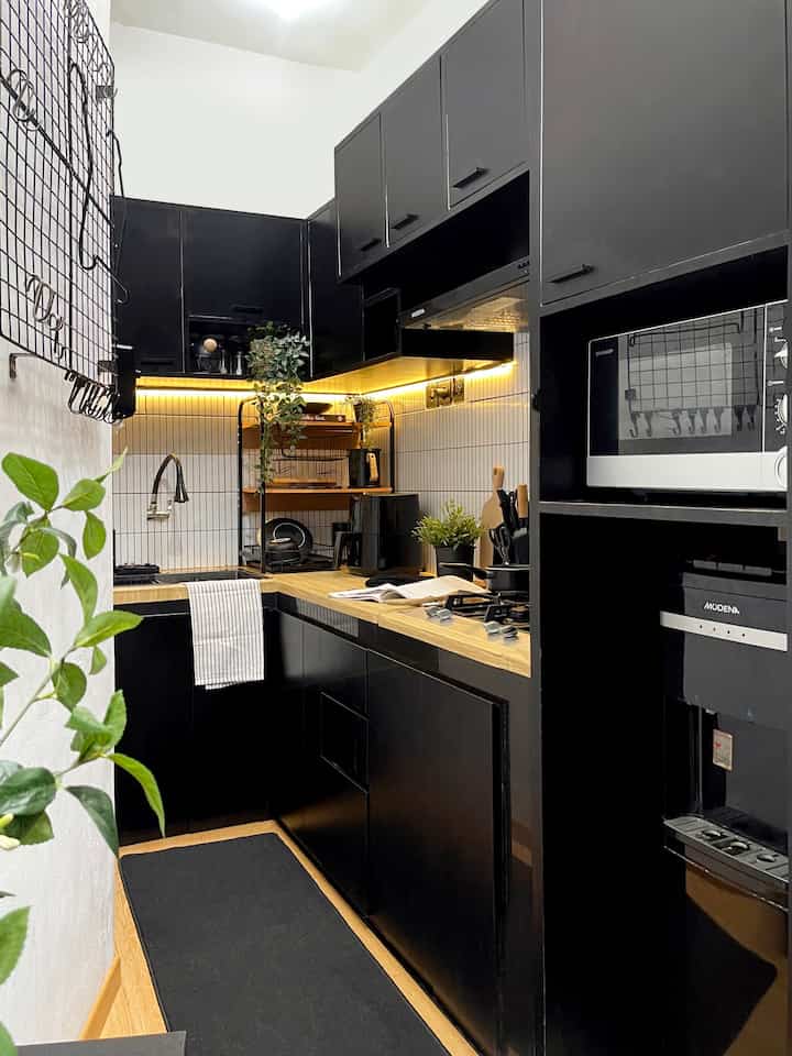 A small L-shaped kitchen in black and wood tones, featuring neat storage and warm under-cabinet lighting.