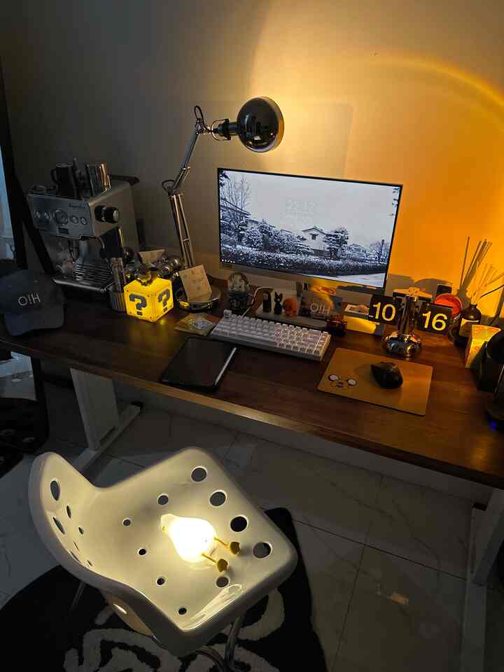 Cozy small home office with brown wooden desk, white office chair, monitor, coffee machine, and LED desk lamp creating a warm workspace atmosphere