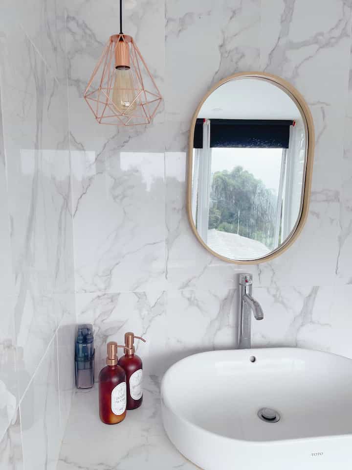 Light gray and white marble tiled bathroom with oval mirror and rose gold pendant light over modern sink area