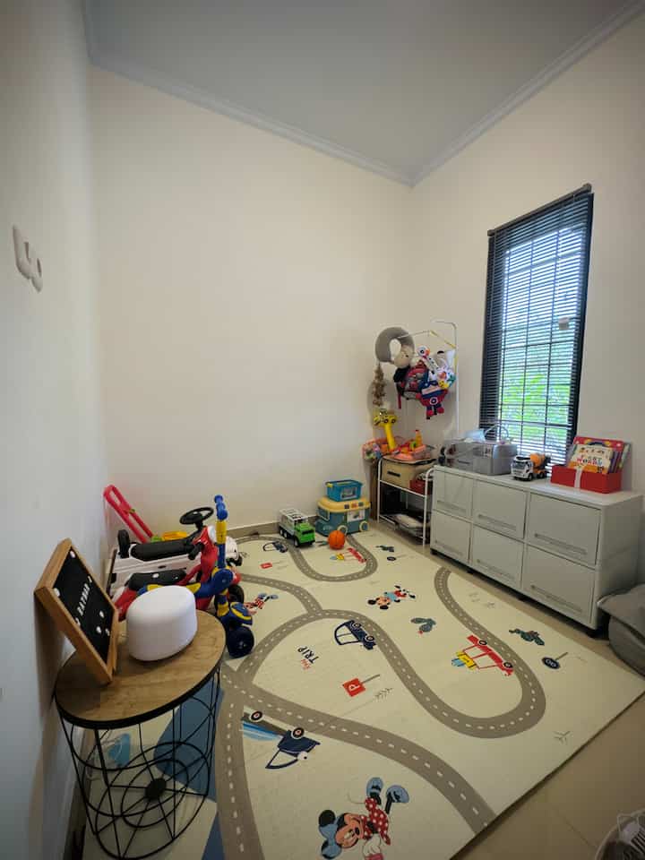 A narrow kids' room with white walls and beige-toned floor featuring a large character playmat, multiple toys, and storage units creating a cozy play area