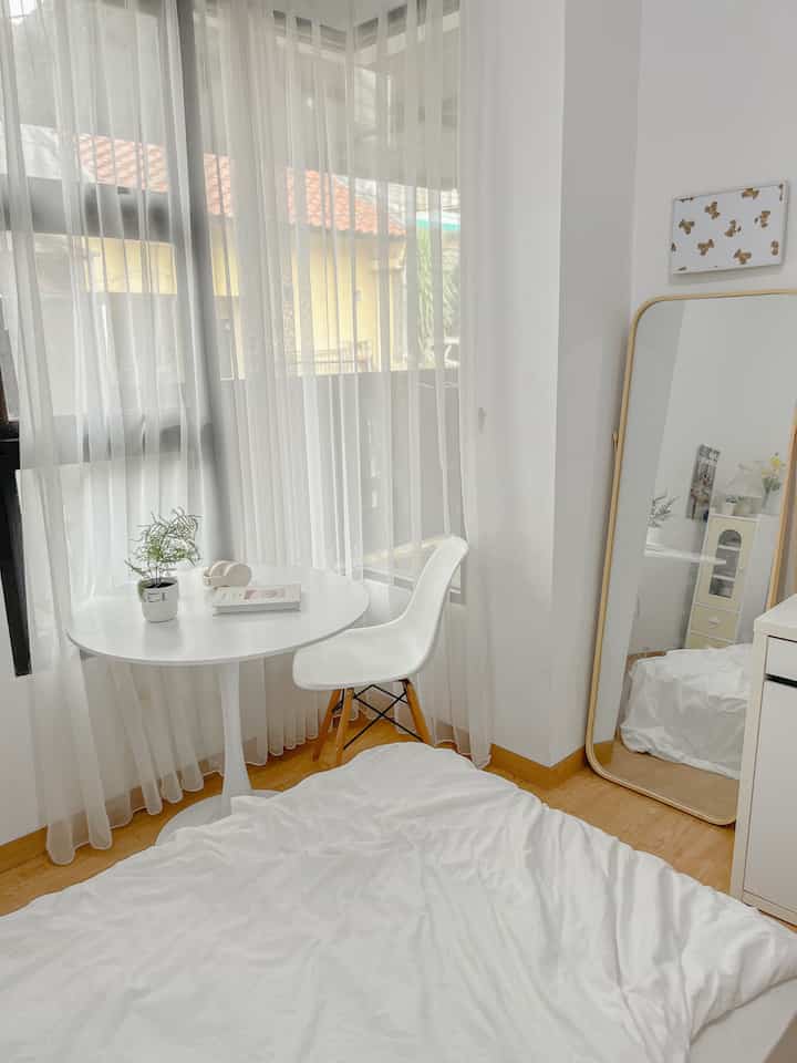 Bright white-toned small bedroom featuring a round table, chair, sheer white curtains, and a small plant for a natural feel