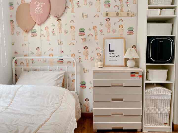 Natural toned kids' room featuring a white metal bed, beige storage drawers, and playful doll wallpaper with a clean, cozy atmosphere