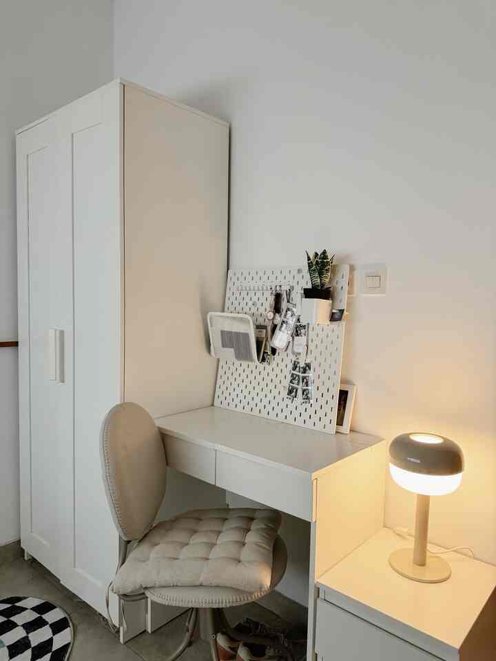 Clean workspace in a studio apartment with white and beige tones featuring a desk, chair, and table lamp