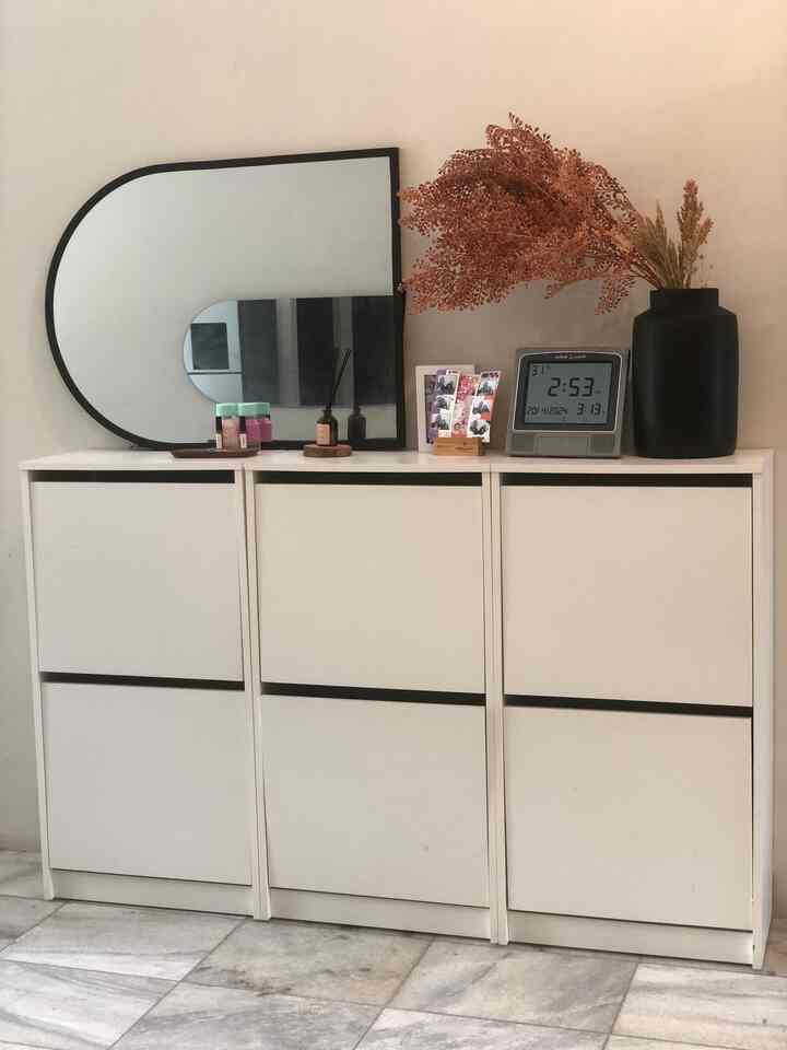 Soft white-toned entrance space featuring a simple white cabinet, large black-framed wall mirror, and brown dried flower decor, showcasing minimalist interior