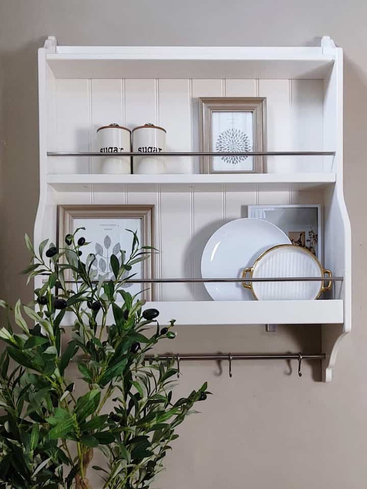 White-toned kitchen wall-mounted shelf featuring framed decorations, dishes, and green plant in a clean and simple space
