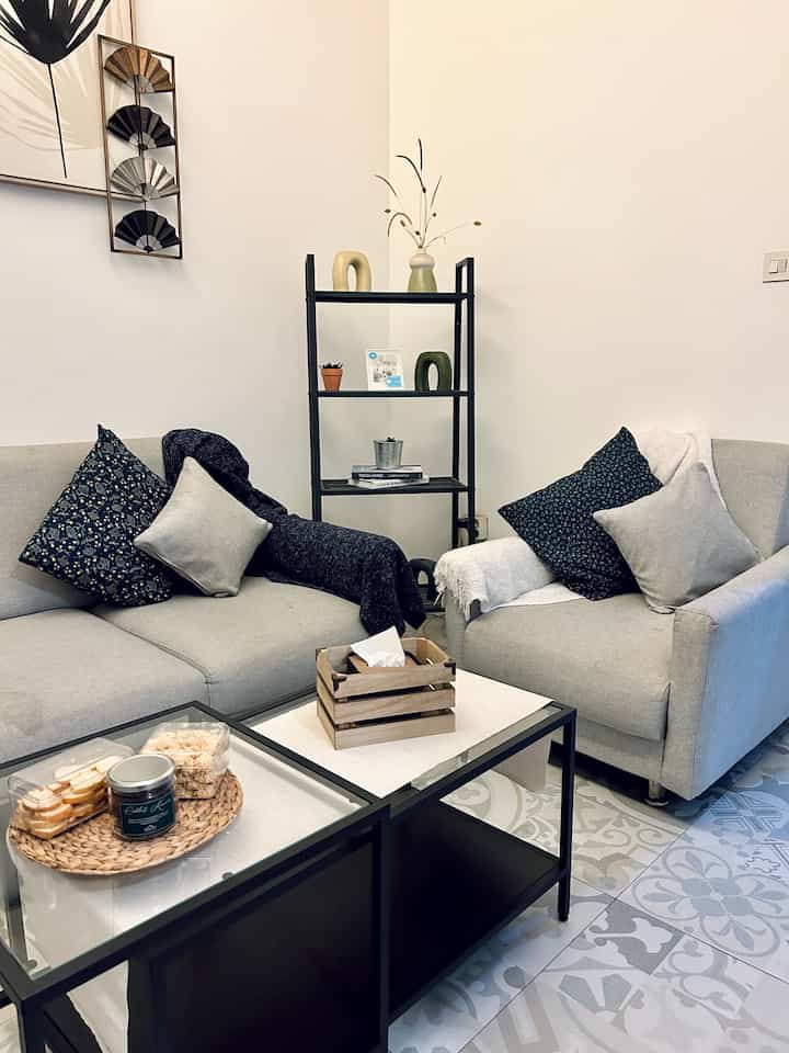 White and gray toned living room featuring modern sofas with cushions and throws, alongside a black metal coffee table and open rack, creating a cozy atmosphere