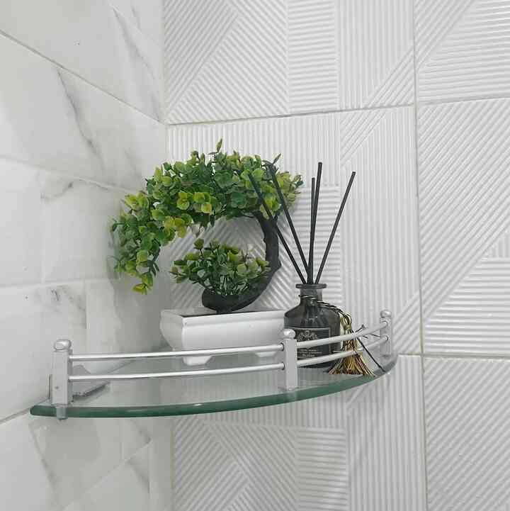 Clean bathroom corner with white tiled walls featuring a glass corner shelf holding a bonsai plant and reed diffuser