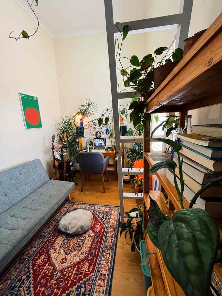 Natural-toned bedroom and home office featuring wooden desk, gray sofa, traditional carpet, and abundant plants creating a cozy urban jungle style room