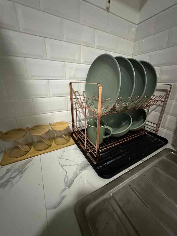 Kitchen corner with white brick tile wall, marble patterned floor, brown metal dish rack holding green plates and bowls, and three transparent glass spice jars