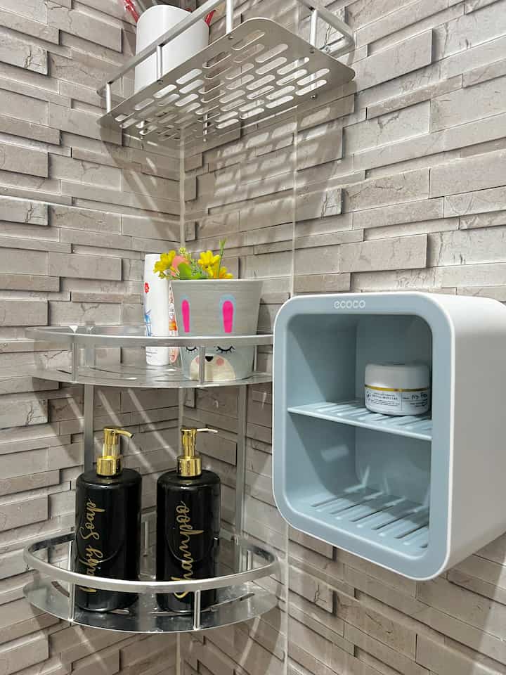 Bright white tiled bathroom corner with metal corner shelves, black and gold pump dispensers, and pastel blue bathroom storage unit