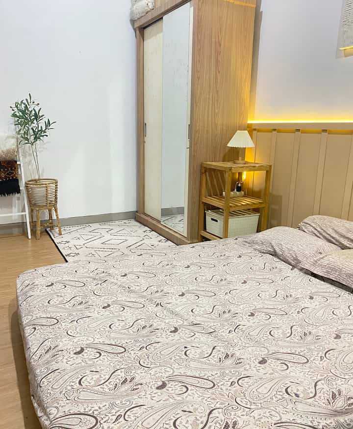 Beige and wood-toned bedroom featuring patterned bedspread, wooden storage shelf, and rattan plant stand creating cozy ambiance