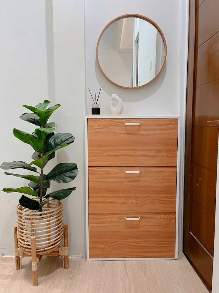 Natural-toned entrance area featuring a white and wood finish shoe cabinet, rattan plant stand with greenery, and a round mirror providing a clean aesthetic