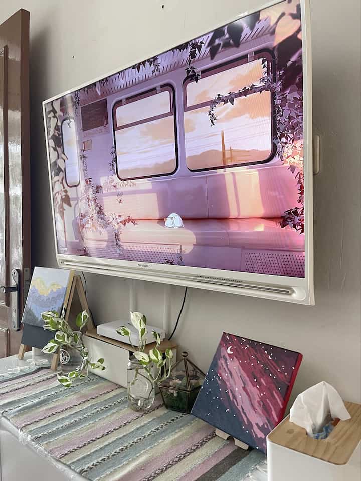 Compact living room in white and wood tones featuring a wall-mounted TV, plants, and picture frames with a simple atmosphere