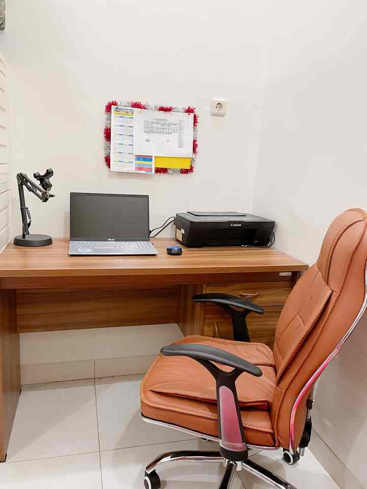 Natural wood tone compact home office with white walls and floor, featuring brown leather office chair, laptop, and printer in a tidy workspace