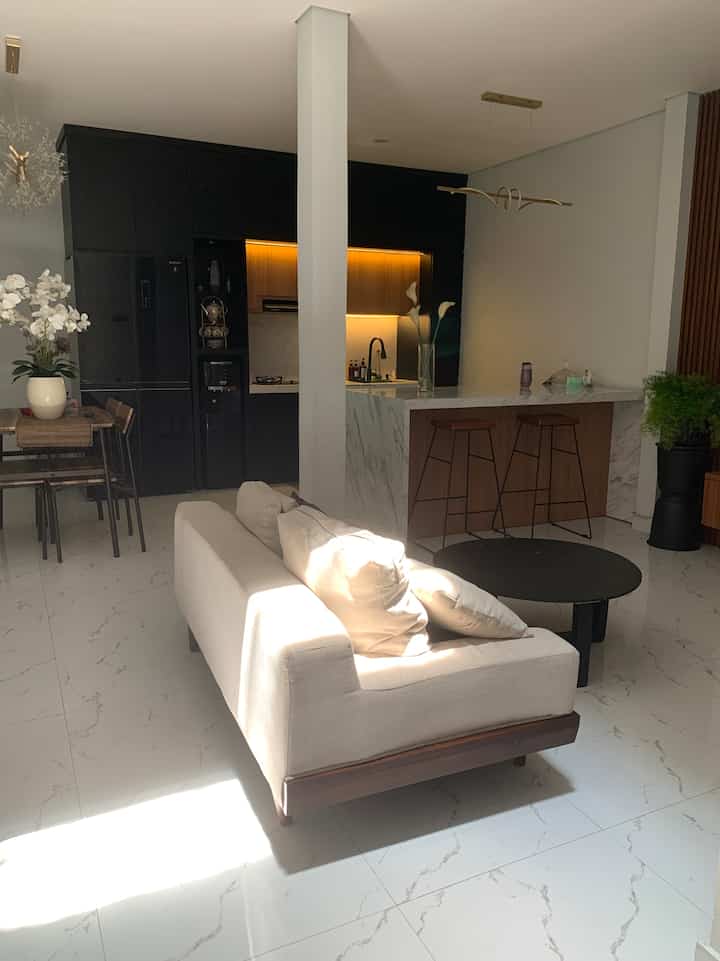A white and black-toned open floor plan living room and kitchen space featuring beige sofa and bar stools, with minimal and natural interior style