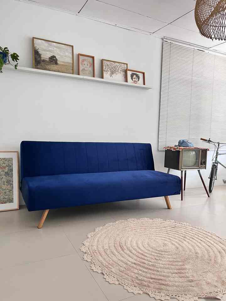 Living room with white walls and blinds, featuring a central blue sofa bed and round beige carpet in a simple style