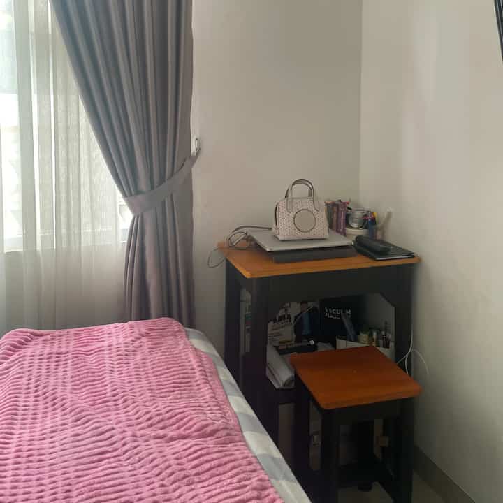 Small bedroom with white walls and beige curtains, featuring a bed with pink bedding and a compact desk with stool, creating a cozy atmosphere