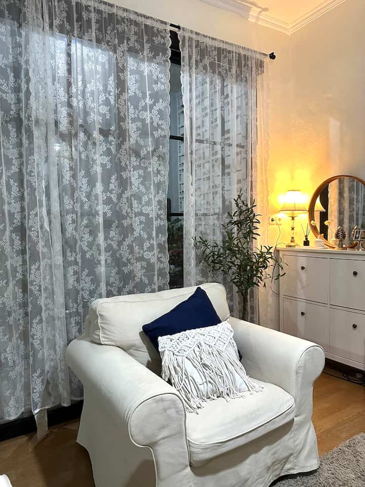 White-toned living room featuring a white armchair, lace curtains, and a sideboard with lighting and mirror creating cozy atmosphere