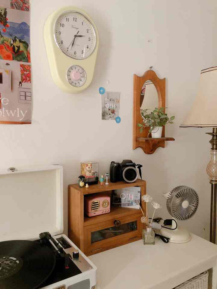 A cozy vintage-style small room corner featuring white walls, wood tone furniture, a clock, table lamp, and various decorative items