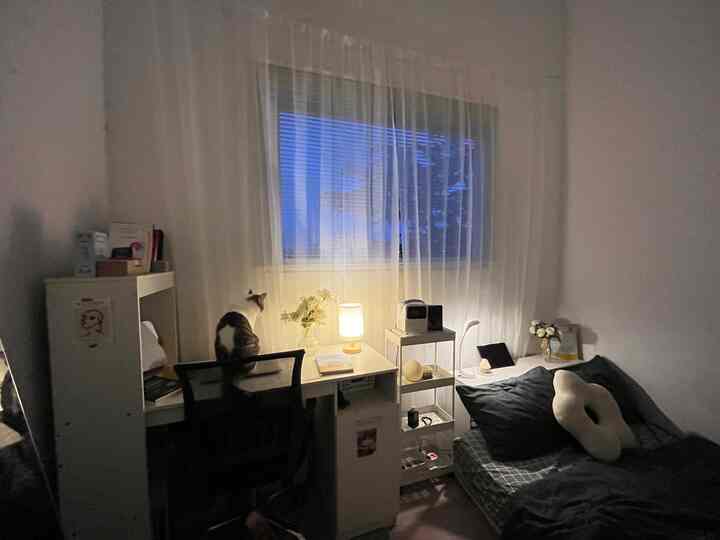 Cozy compact bedroom with warm lighting, white desk and curtains, and a cat sitting on the chair