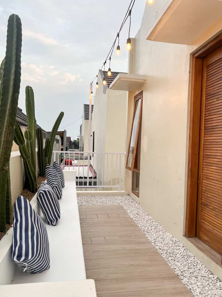 Bright, natural balcony featuring white bench seating, brown wood-tone elements, cacti plants, and string lights above