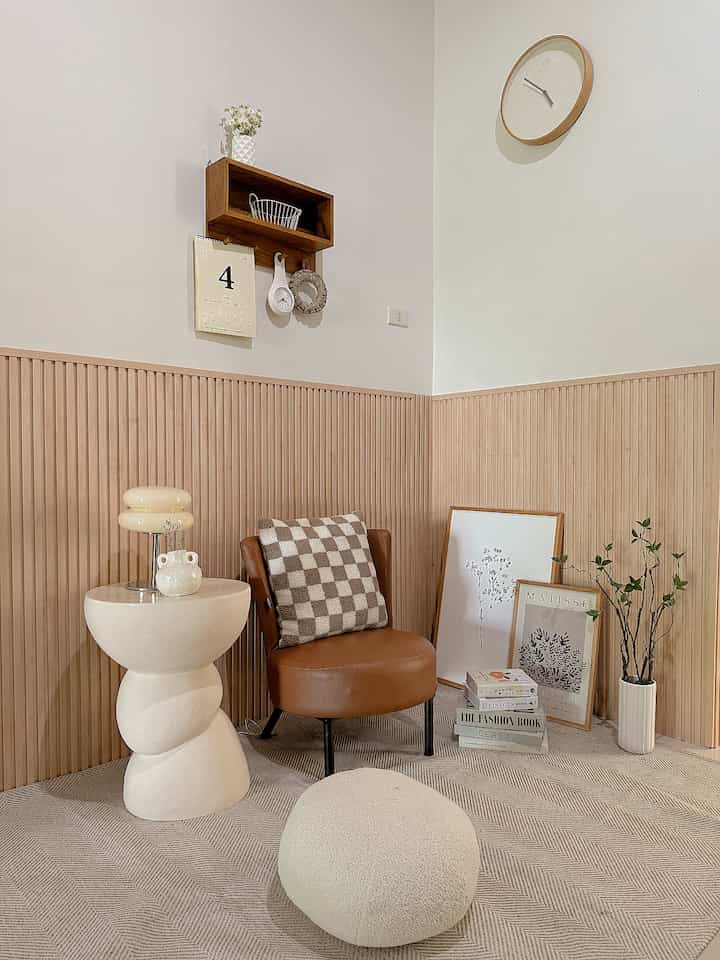 Natural tone living room corner featuring a brown armchair, unique cream side table, wall clock, and plant creating a cozy atmosphere