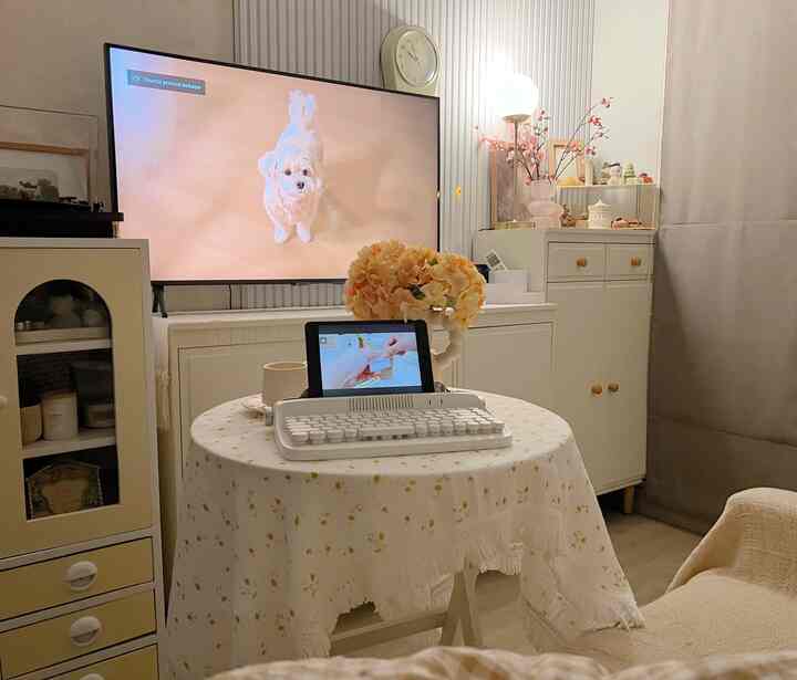 Small living room and workspace in white and beige tones featuring a folding table with keyboard and tablet, TV and wall clock mounted on the wall, cozy vintage style interior