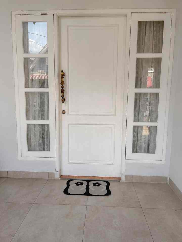 White-toned entrance area featuring a central white door with side windows and a flower-patterned carpet placed in front of the door, creating a neat space