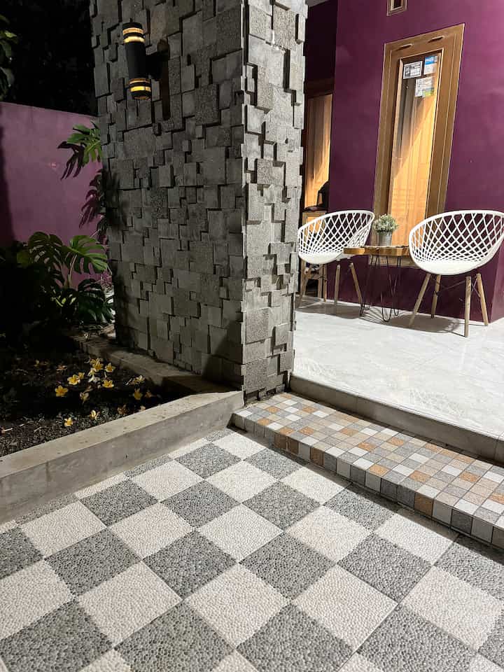 Gray and white tiled floor balcony space with purple walls, featuring two white rattan chairs and wooden table in a serene outdoor setting