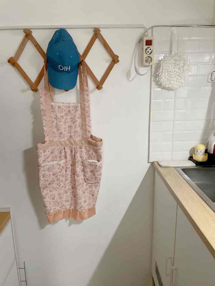 A kitchen corner with white walls and natural wood tones, featuring a pink floral apron and blue cap hanging on a wooden rack