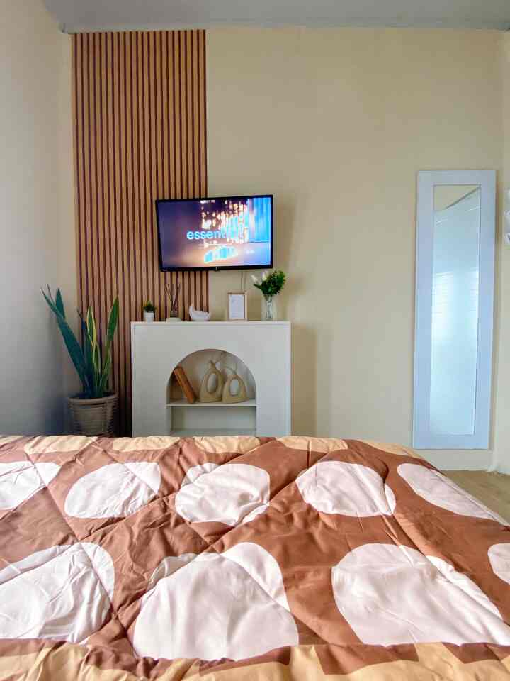 Beige and brown-toned bedroom featuring a bed in the center, wooden paneling on left wall, full-length mirror on right, and a mounted TV, creating a simple cozy interior