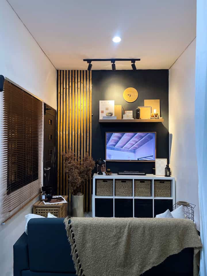 Natural modern small room living space with dark accent wall and wood tones, featuring bookshelf, sofa, and blinds