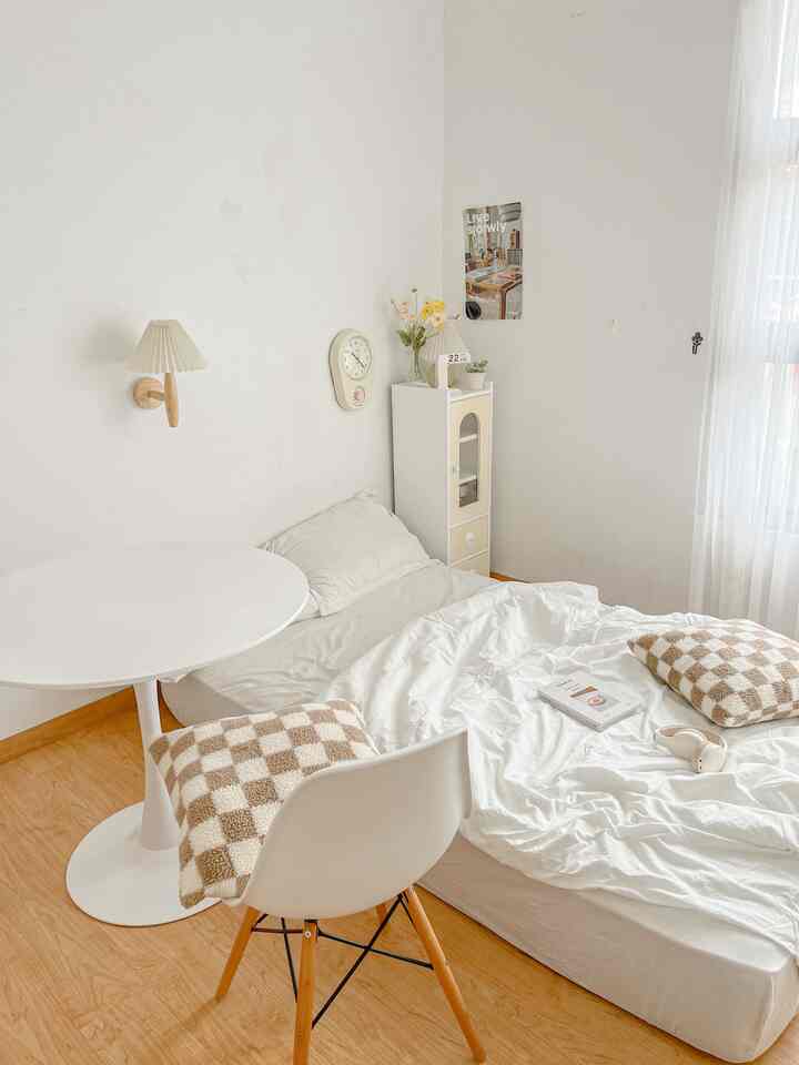 A simple bedroom in white and wood tones featuring a white bedspread and round table, creating a cozy atmosphere