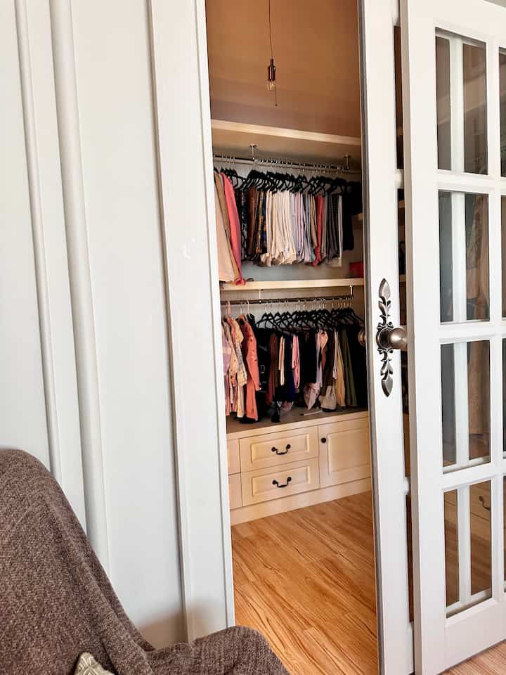 A white and brown toned walk-in closet entrance with organized wardrobes and a cozy armchair