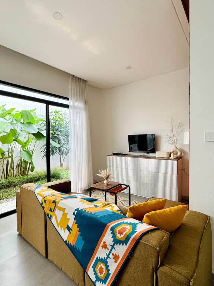 Beige-toned compact living room featuring a large sofa, TV stand, sheer curtains, and green plants in a modern setting