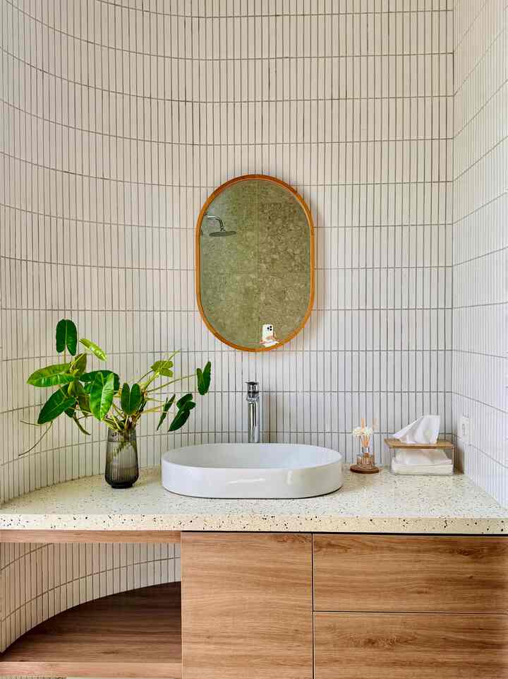 White tiled bathroom wash basin with wood tone cabinetry, oval mirror and green plant arranged in a clean setting