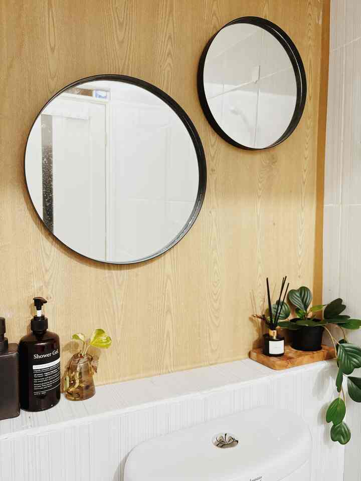 White tiled bathroom with wood tone wall, featuring round mirrors, diffuser, and plants in a clean minimalist space