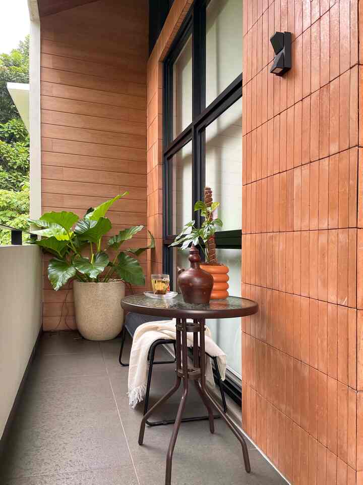 Natural tones and brown terracotta walls enclose a narrow, long balcony featuring a table, chairs, and tropical plants creating a cozy atmosphere