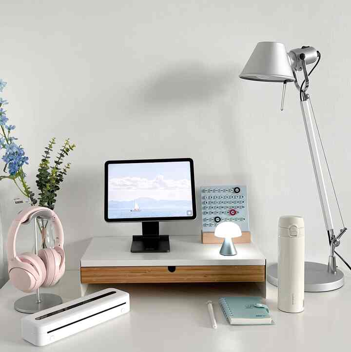 White and silver toned minimal home office space featuring a desk with tablet, headphones, and lighting in a clean workspace
