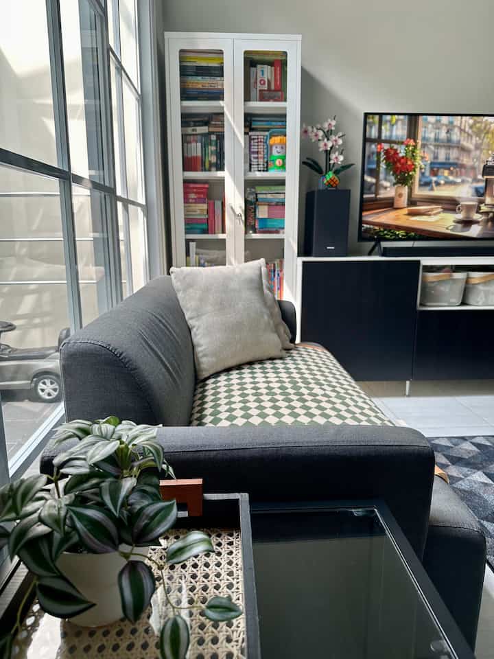 A black and white toned living room featuring a sofa, bookshelf, and TV stand with calm ambiance and ample natural light