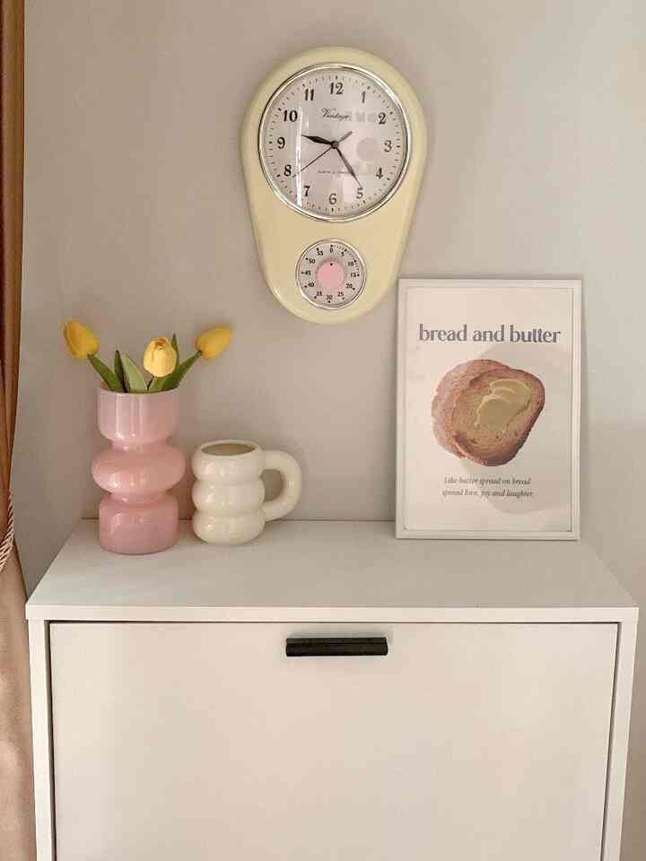 A simple interior space with beige walls and white cabinet featuring pink vase with yellow tulips, vintage wall clock, and bread-themed framed art