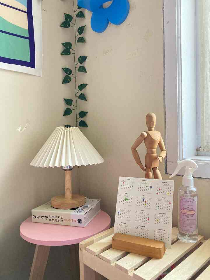 Soft white and pink toned small corner space featuring a table lamp, calendar, and wooden mannequin with a minimal atmosphere