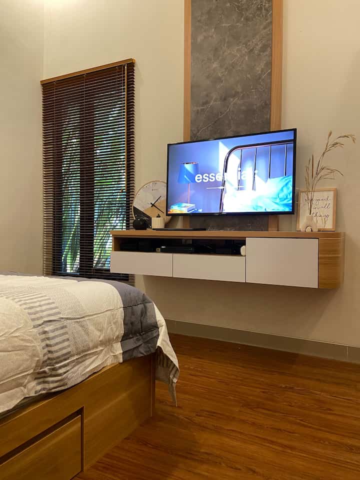 Warm modern bedroom featuring wood-tone flooring and bed, wall-mounted TV, and wooden blinds