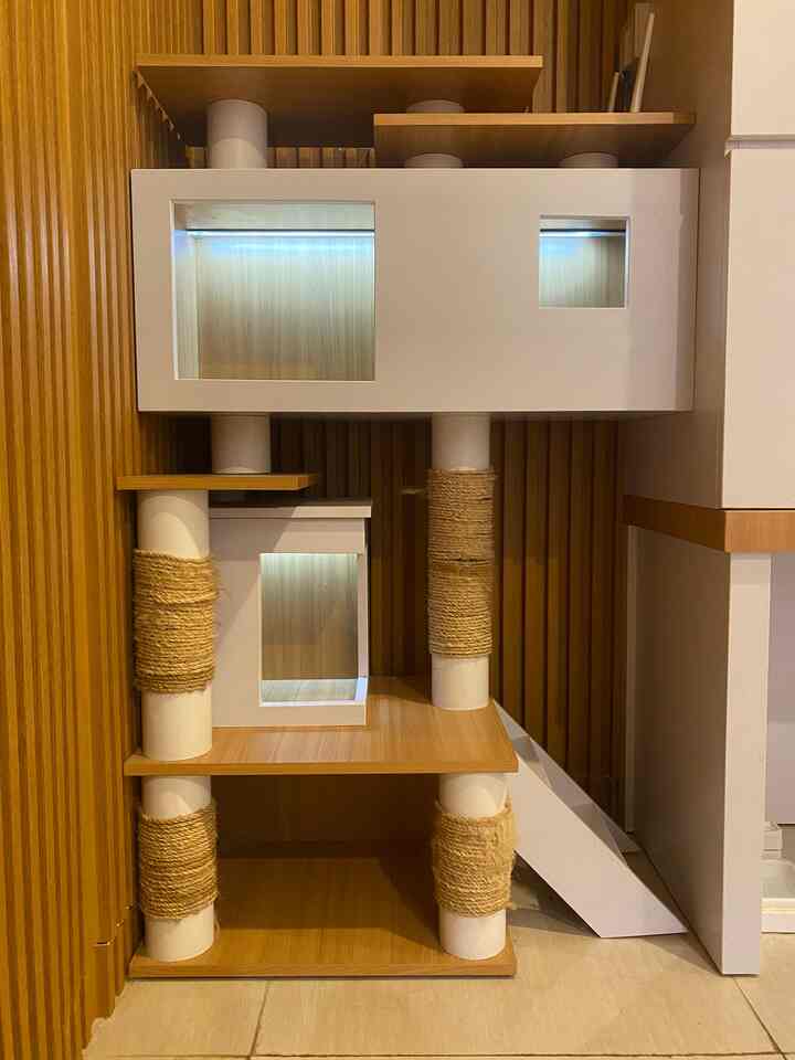 A white and wood-toned multi-level cat tower is placed against a wood-paneled wall, creating a cozy pet area
